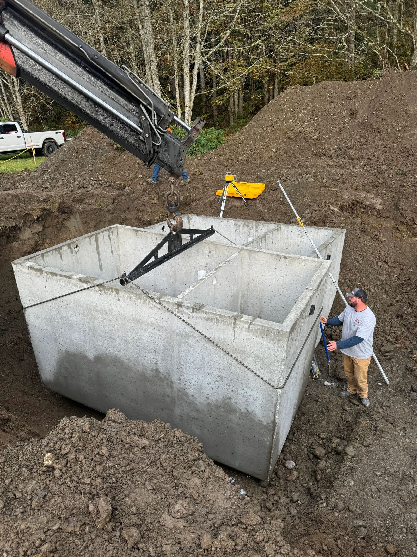 A concrete septic tank being craned into an excavation while a technician guides it.
